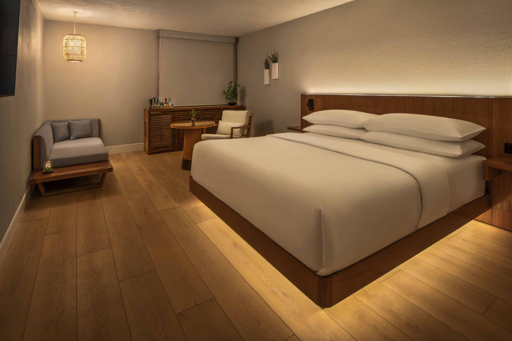A modern hotel room with a large wooden bed, ambient under-bed lighting, a seating area, desk, and warm wood tones.