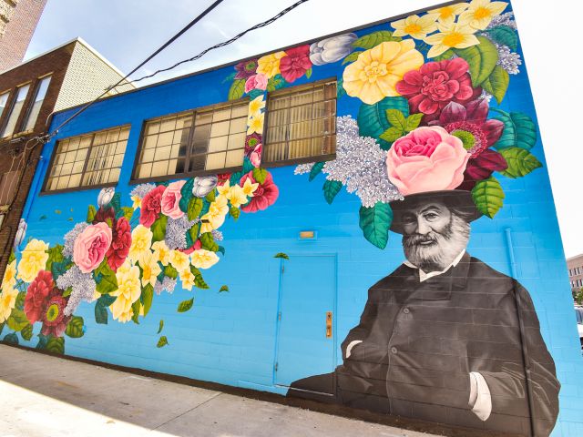 The image shows a mural on a brick wall with a figure and colorful floral elements, lit by string lights in an alleyway at night.
