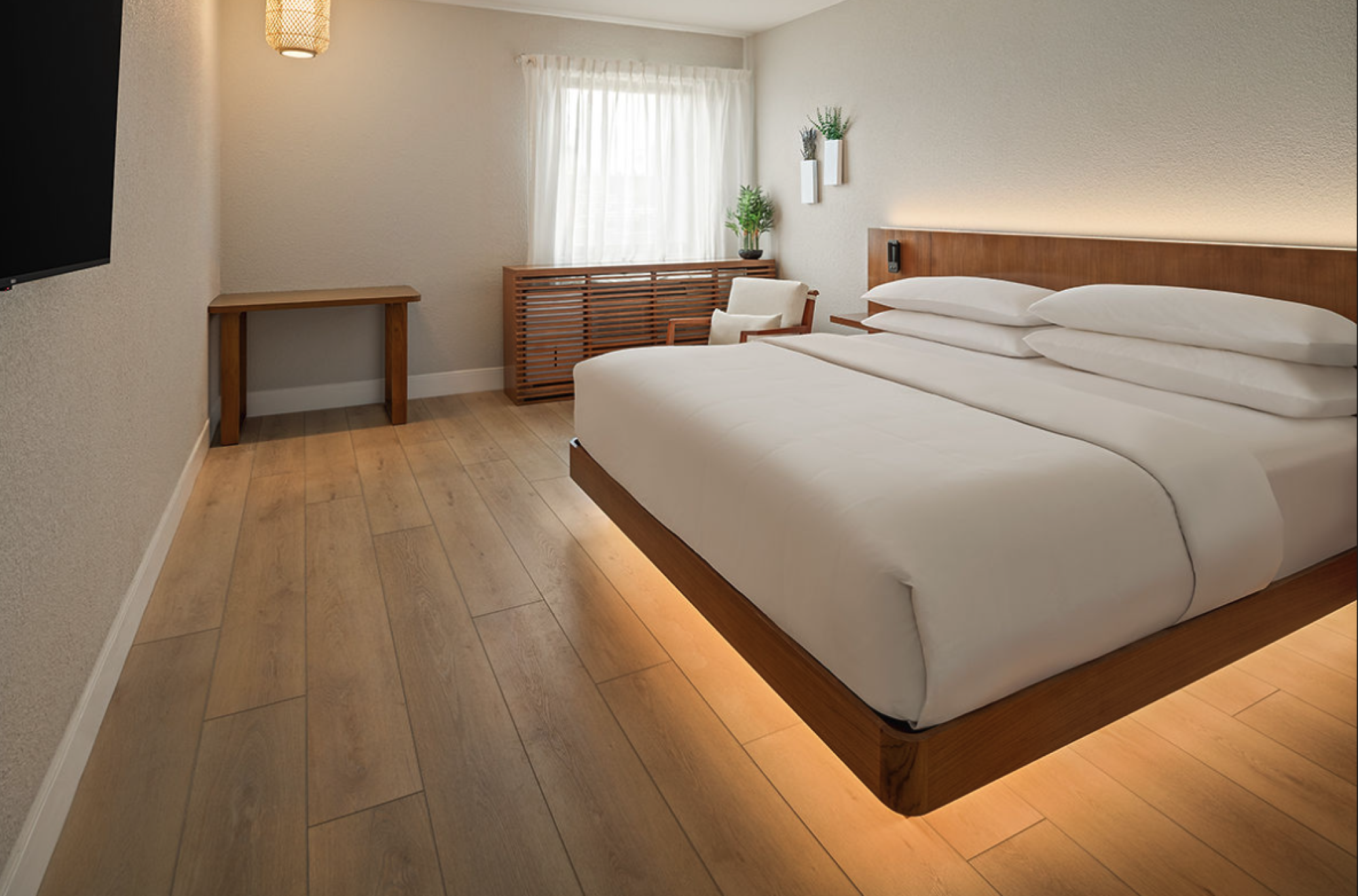 A modern hotel room with a low wood bed, white linens, warm lighting under the bed, a wooden desk, dresser, chair, and a window with sheer curtains.