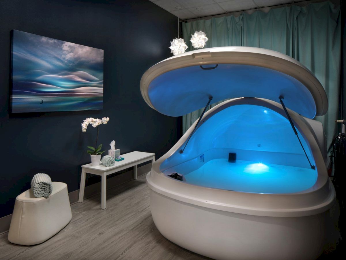 A room with a sensory deprivation tank, ambient blue lighting, a decorative table, wall art, orchid, and a small decorative item.