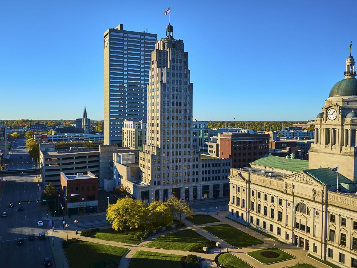 A cityscape featuring a mix of historic and modern buildings under a clear blue sky with green trees lining the streets.