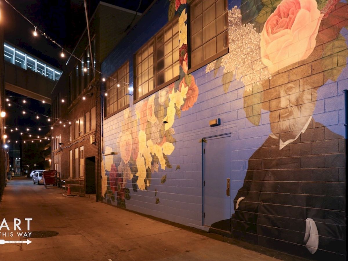 A lit alleyway with a colorful mural featuring a man in a suit with flowers and plants around his head. String lights adorn the area.