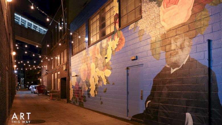 A lit alleyway with a colorful mural featuring a man in a suit with flowers and plants around his head. String lights adorn the area.