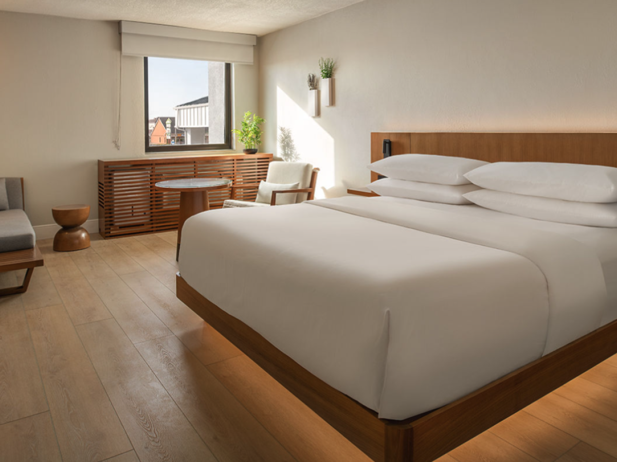 A stylish hotel room with a large bed, warm wood tones, a seating area, and soft ambient lighting near a window overlooking a cityscape.