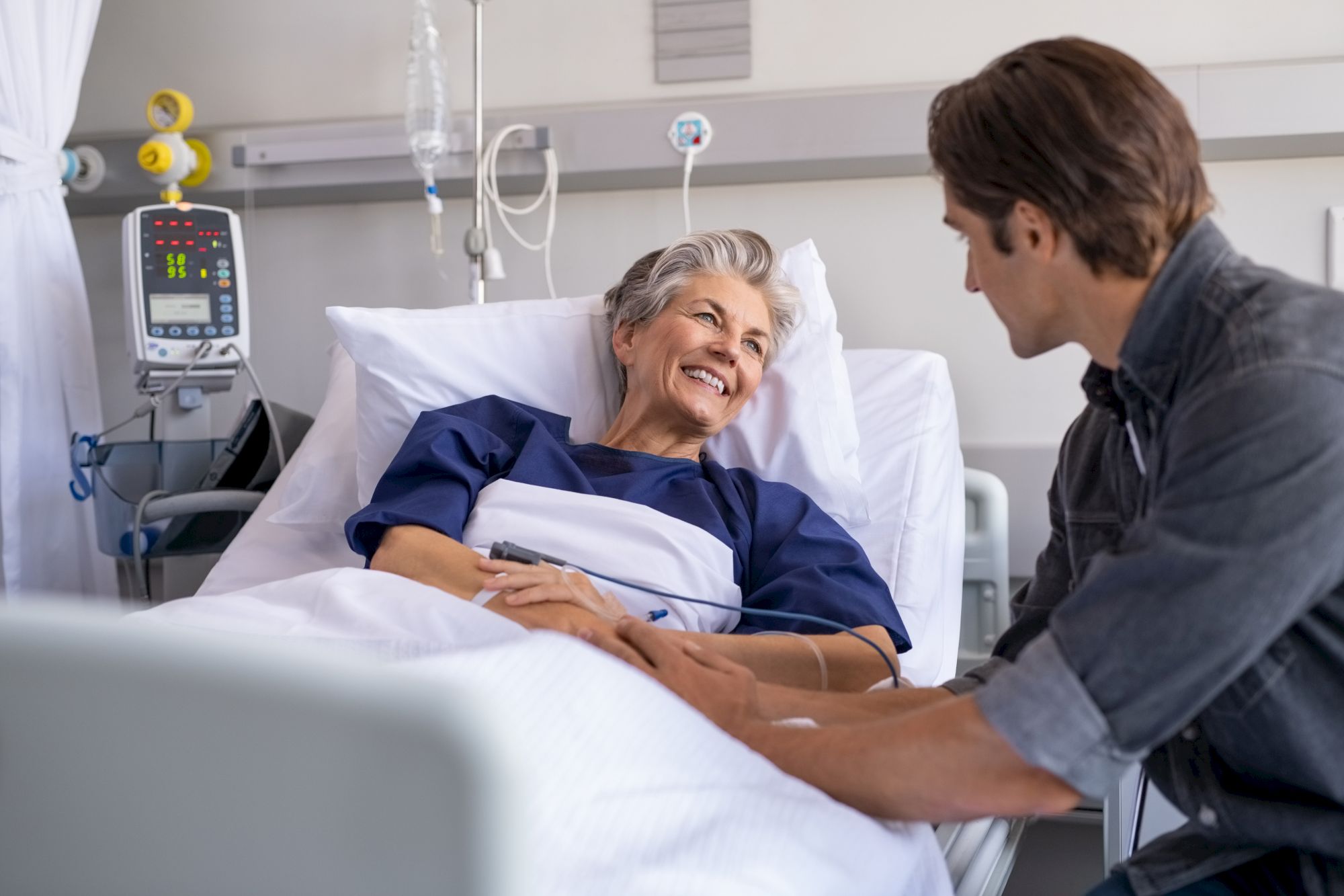 An elderly patient in a hospital bed smiles while a visitor holds her hand, creating a warm and caring atmosphere in the room.