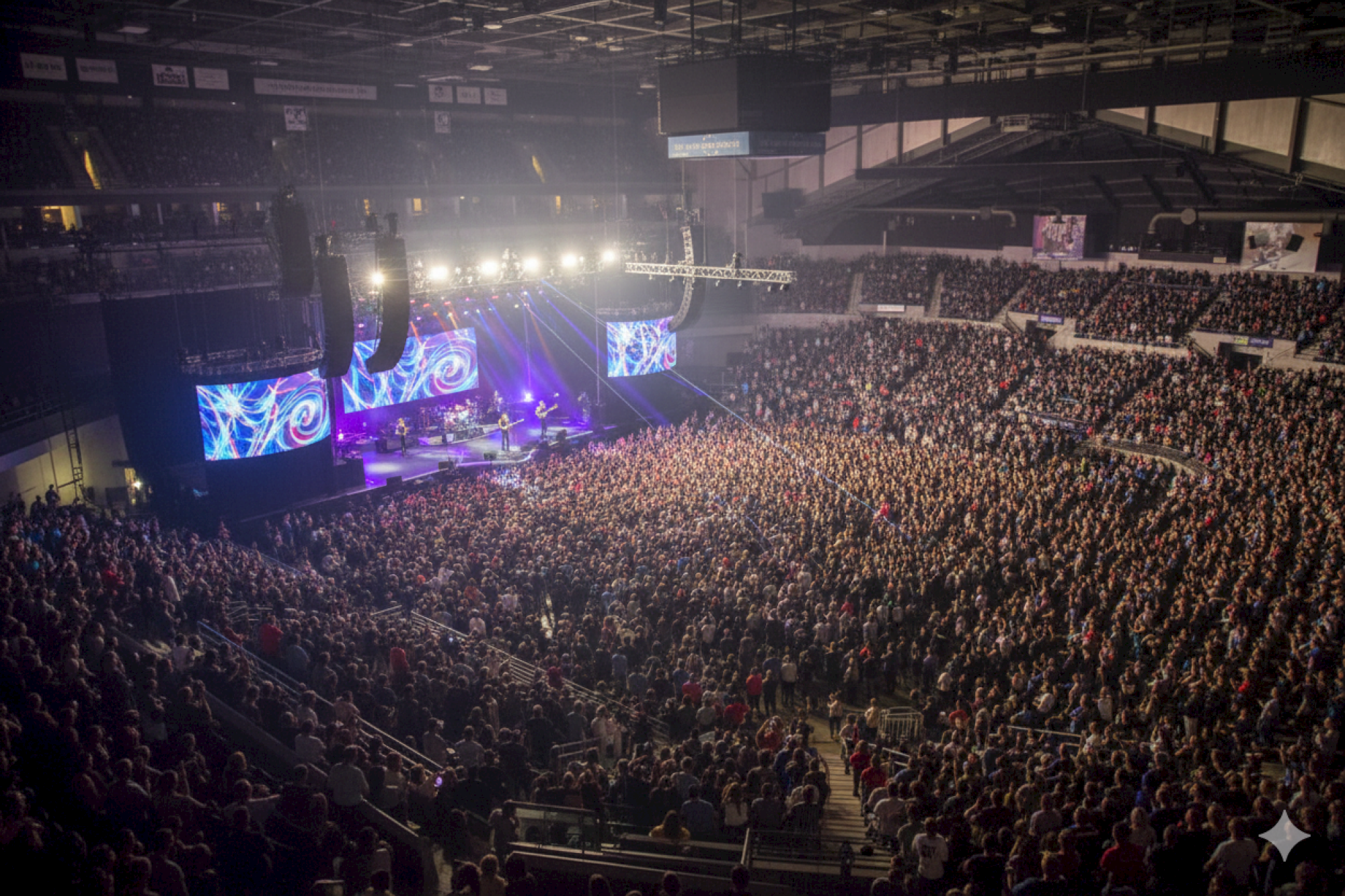 A large crowd is gathered at an indoor concert, with bright stage lights and screens displaying visuals.